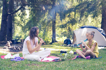 Mom and daughter are playing on the flutes, sitting in a forest glade