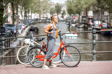 Young beautiful woman riding a bicycle on the bridge over the water channel in Amsterdam old city - obrazy, fototapety, plakaty