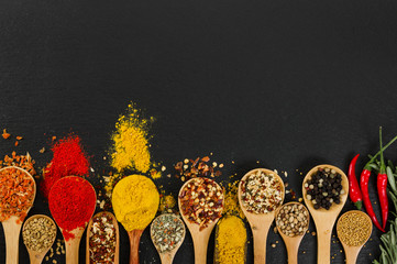 Various spices in wooden spoons on black slate background.