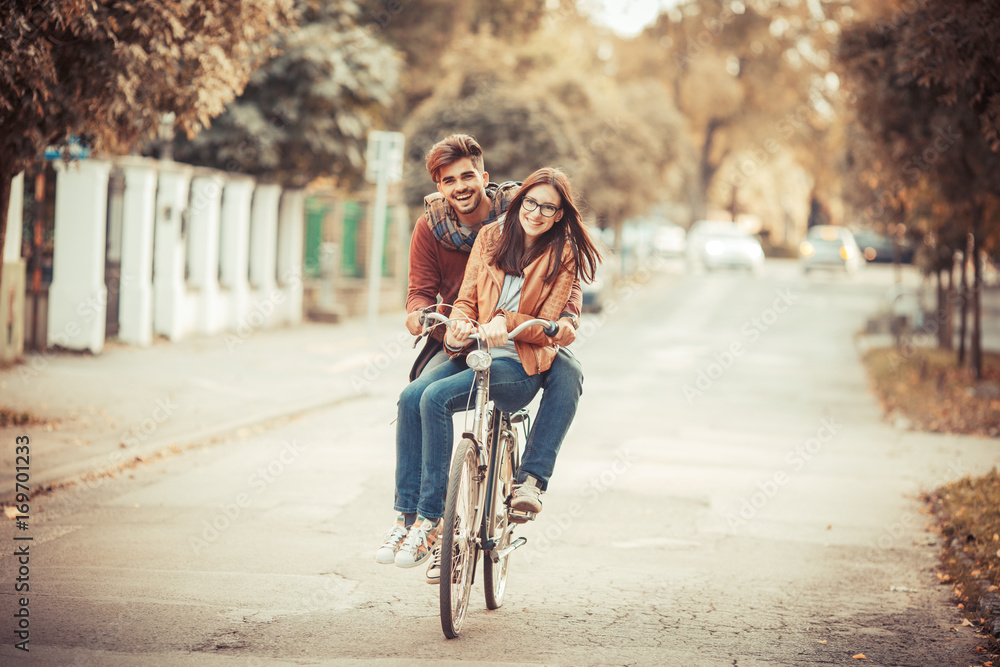 Wall mural Young couple riding bicycle at the street on autumn day.They sitting on bike and making fun.