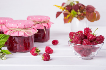 Raspberry jam in glass jar, fresh  ripe raspberry and green leaves on white wooden table
