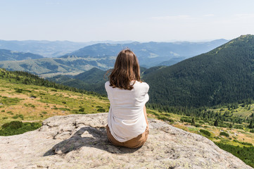 Naklejka premium Back of young female person sitting on top of the rock in ukrainian carpathian mountains. Girl tourist enjoys gorgeous landscape scenery on hot sunny day