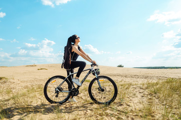 Naklejka premium Woman cyclist carrying mountain bike on sunrise trail. Girl in sport wear on black white bicycle going to record in summer sunset time in desert.