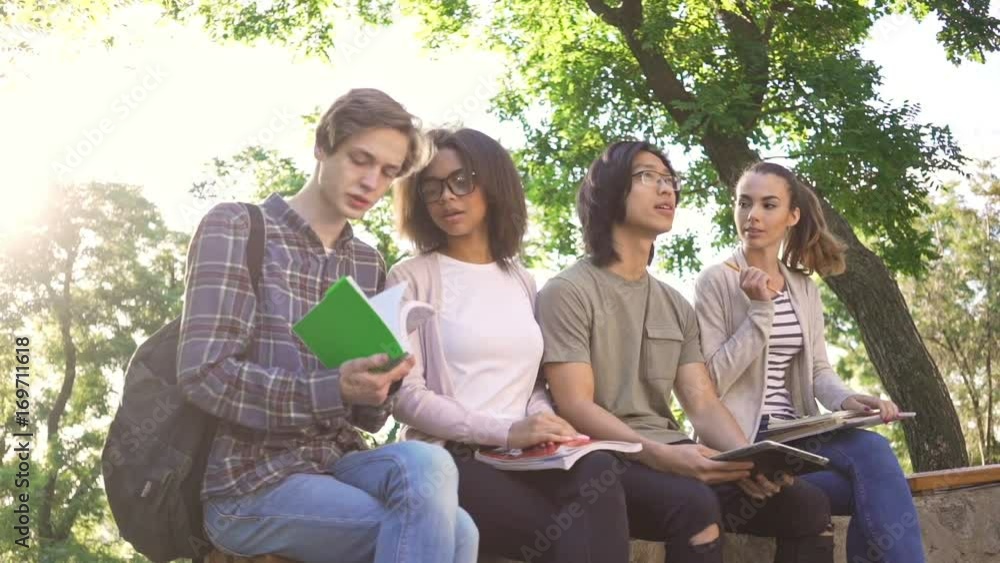 Poster Multiethnic group of cheerful young students sitting and talking outdoors while studying. Looking aside.