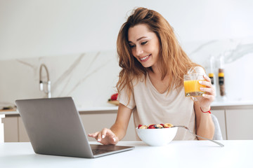 Positive woman with juice using laptop in the kitchen