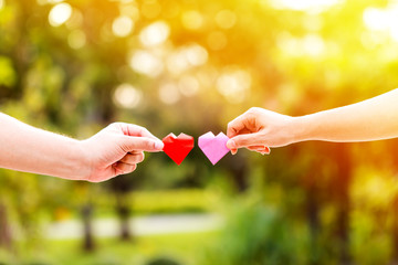 A man and woman hand holding a red heart and pink heart with love in sunlight in the public park.