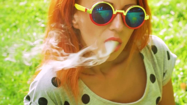 Cute girl with red hair and sunglass at the green grass park and smokes vape and relax close-up with sunlight