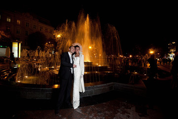 Happu groom and elegant bride stand before the sparkling fountain