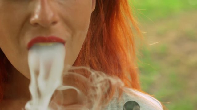 Cute girl with red hair smokes vape in summer park close-up