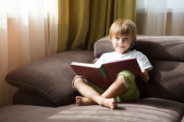 Cute little boy is reading book sitting on a sofa at home