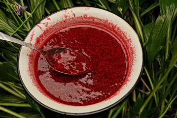 белая миска с малиновым вареньем и ложкой стоящая в зелёных листьях