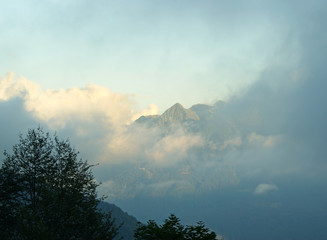 Landscape: wooded mountain peaks, partially covered by clouds and illuminated by the sun