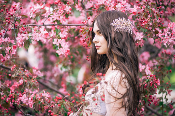 Fototapeta premium piękna śliczna panna młoda brunetka w przezroczystej sukience z buduaru, z dekoracją na włosach, w pobliżu kwiatów drzewa z różowymi kwiatami, profil, poziomy, ogród