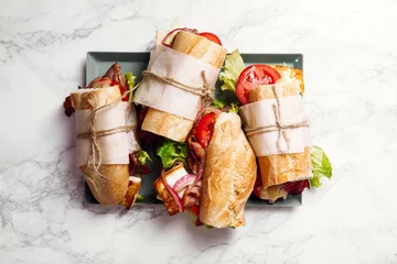 Foto auf Acrylglas Frisches Baguette-Sandwich im Bahn-Mi-Stil. Speck, gerösteter Käse, Tomaten und Salat auf metallischem Tablett auf weißem Marmorhintergrund. © Anastasiia Nurullina
