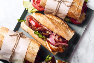 Fresh baguette sandwich bahn-mi styled. Bacon, roasted cheese, tomatoes and lettuce on metallic tray on white marble background. Close up view.