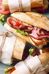 Fresh baguette sandwich bahn-mi styled. Bacon, roasted cheese, tomatoes and lettuce on metallic tray on white marble background. Close up view.