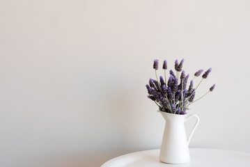 Purple lavender in small white jug on edge of round table against neutral wall with copy space to left