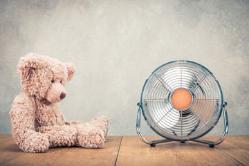 Fototapeta Retro Teddy Bear toy sitting near office or home cooling fan on wooden table front concrete wall background. Vintage old style filtered photo obraz
