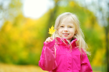 Cute little girl having fun on beautiful autumn day. Happy child playing in autumn park. Kid gathering yellow fall foliage.