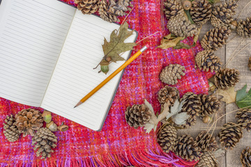 Pencil and open notebook with blank lined pages lying on cozy autumn knit blanket, acorns, leaves and pine cones on wooden background