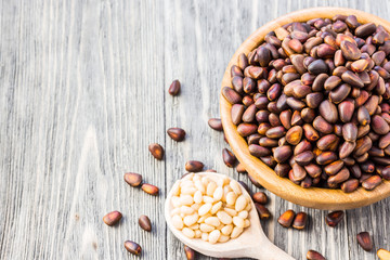 Whole nuts in a bowl and peeled pine nuts in a spoon on wooden background. Selective focus, close up. 