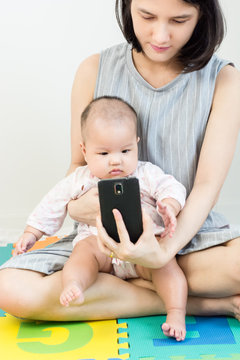 Portrait of adorable baby sitting on mother lap on colorful eva foam and llooking to smart phone.