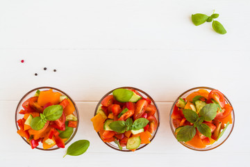 Vegetarian vegetable salad with tomatoes, cucumber, red bell pepper and basil in the glass bowls on the white wooden table, top view.