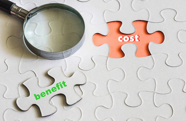 conceptual image of cost and benefit analysis. Magnitying glass on off-white jigsaw puzzle and text cost benefit. Selective focus at center.