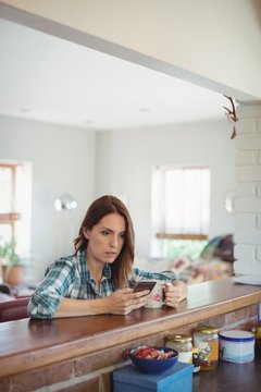 Woman using mobile phone while having coffee in kitchen