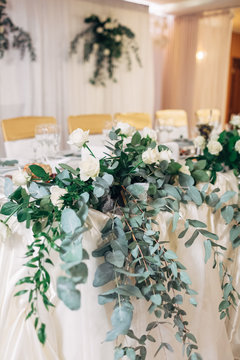 Garland of long green branches and white roses lies on white dinner table