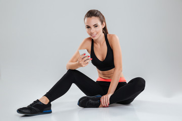 Smiling fitness woman sitting on the floor with smartphone