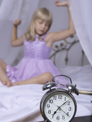 Obraz na płótnie Canvas Alarm clock standing on bedside table. Wake up of an asleep young girl is stretching in bed in background