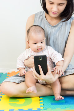 Portrait of adorable baby sitting on mother lap on colorful eva foam and llooking to smart phone.