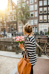 Fototapeta premium Lifestyle portrait of a woman walking with bouquet of pink tulips near the water channel in Amsterdam city