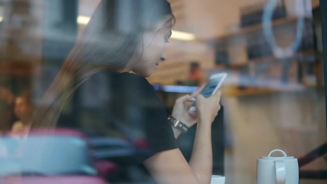 View from the outside of a concentrated young lady with long hair messaging using her mobile phone in the coffee shop. White cup with saucer and teapot on the wooden table