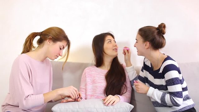 Lovely women spend time together and engaged in women's affairs. Two women help to get girlfriend to meet with guy, paint nails and lips, braid hair, chat and joke, sitting on soft gray sofa in bright