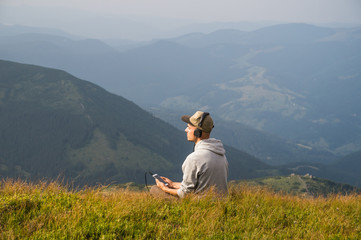 Fototapeta na wymiar Man sits top of the hill in carpathian mountains and listens to music in headphones and enjoys beautiful view. Young male person enjoys music in headset from cell phone on a mountain meadow