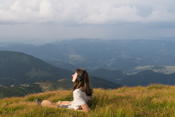 Young woman takes rest top of the hill in carpathian mountains and listens music in headphones enjoying beautiful scenery. Female hiker in headset with cell phone on a mountain meadow