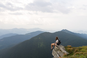 Young female hiker sits on rock in gorgeous mountain area and looks in endless horizon. Woman with tourist rusksack takes rest top of hill after reaching destination in carpathians  