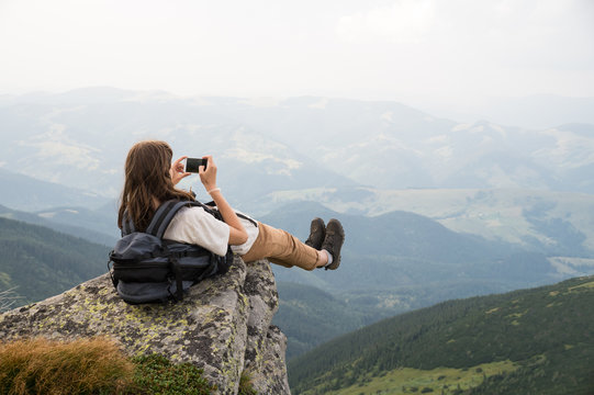 Female tourist takes photo of legs with cell phone from top of the mountain cliff. Hiker girl with backpack taking self portrait in beautiful scenery on a rock in ukrainian carpathians