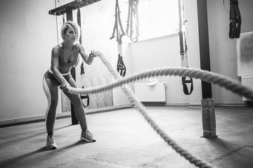Fit blonde beautiful woman with battle ropes exercise in the fitness gym. Young female wearing sportswear.