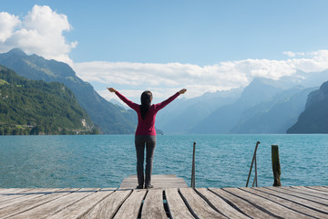 Woman with open arms standing on the shore of the lake. Mountain range in the background.