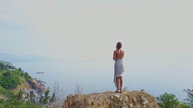 Blond Girl Stands on Rock against Green Hill and Ocean