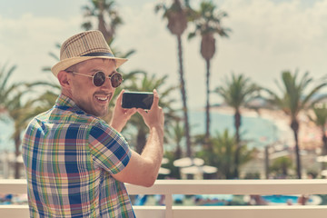 Man in sunhat looking in the camera while making photo of amazing sea view with his smartphone. Santa Susanna. Cataluna. Spain.