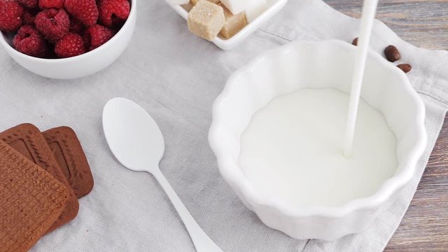 Healthy breakfast - pouring milk, yogurt into white bowl, adding cereals and berries. Top view on table with chocolate cakes, healthy super bowl food, raspberry and white dishes. Camera locked down.