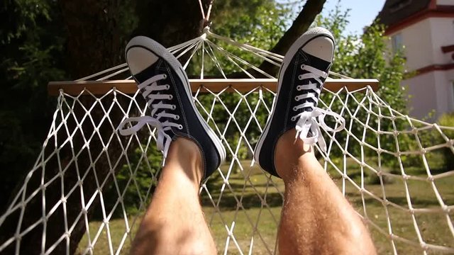 A man relaxing in a hammock in the garden. Male legs in sneakers. Chilling out in summertime. Point of view. 