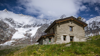 Close up view of traditional rural house in the Pennine Alps. Green grass around, blue sky with white clouds above and mountains behind house. Summer in the Pennine Alps, Valle d'Aosta, Italy, Europe.