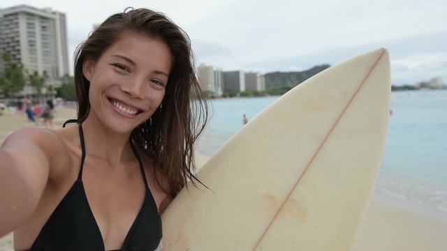 Selfie video - Surfing surfer girl taking self portrait photo holding surfboard on beach. Sexy beautiful bikini woman living healthy active water sports lifestyle. Asian model, Waikiki, Oahu, Hawaii.