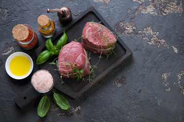 Black wooden serving board with raw medallion beefsteaks and condiments, high angle view with space, brown stone background
