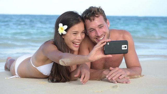 Selfie couple beach holiday taking with smartphone lying down relaxing and having fun holding smart phone camera. Young beautiful multicultural Asian Caucasian couple having fun on summer beach.